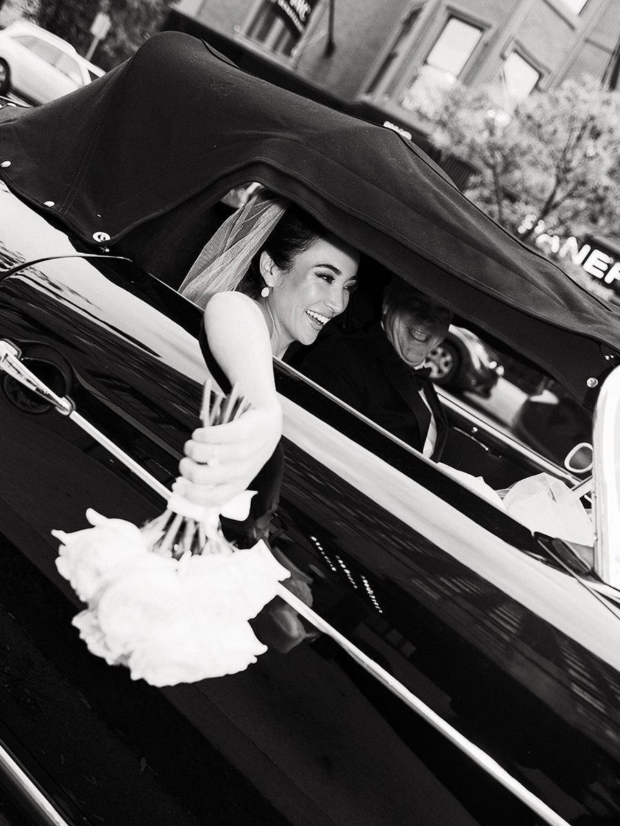 A smiling bride wearing a veil is sitting in a classic convertible car with the top up, holding a bouquet of flowers out of the window. A man, likely the groom, is seated next to her, both dressed formally. The photo is in black and white.