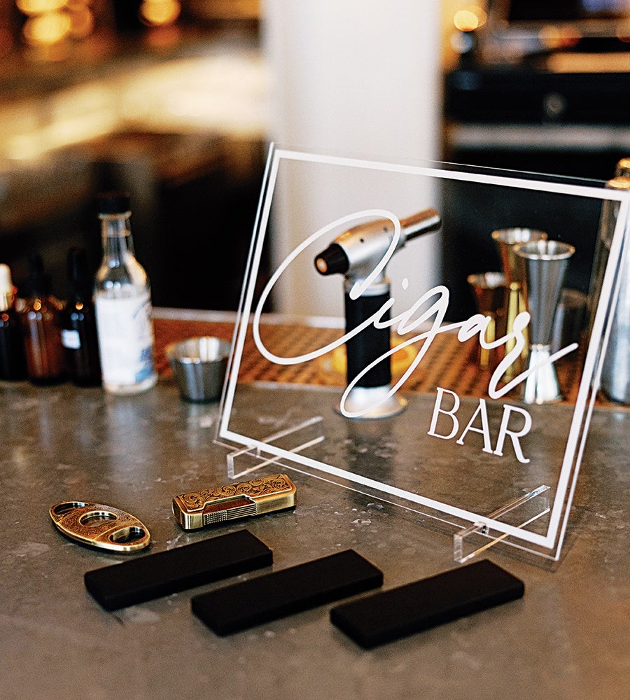 A clear acrylic sign with white cursive and block lettering reading "Cigars BAR" is displayed on a countertop. In front of the sign are a gold cigar cutter, a gold lighter with intricate designs, and three black rectangular objects. In the background, there is a small torch lighter and various bottles and metal jiggers.