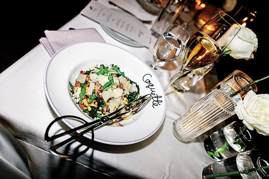A white plate with the word "Coquette" contains a salad topped with shaved cheese and fresh herbs. The plate is on a white tablecloth next to a folded pink napkin. Nearby are a pair of silver tongs, a glass of champagne, a glass of water, and two white roses in small vases. A menu and additional glassware are visible in the background.