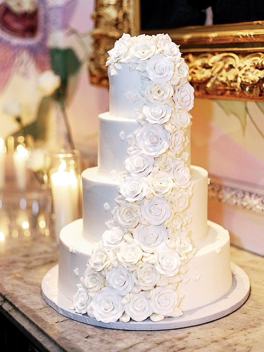 Four-tier white wedding cake decorated with cascading white fondant roses, placed on a marble surface with lit candles and a gold-framed painting in the background.