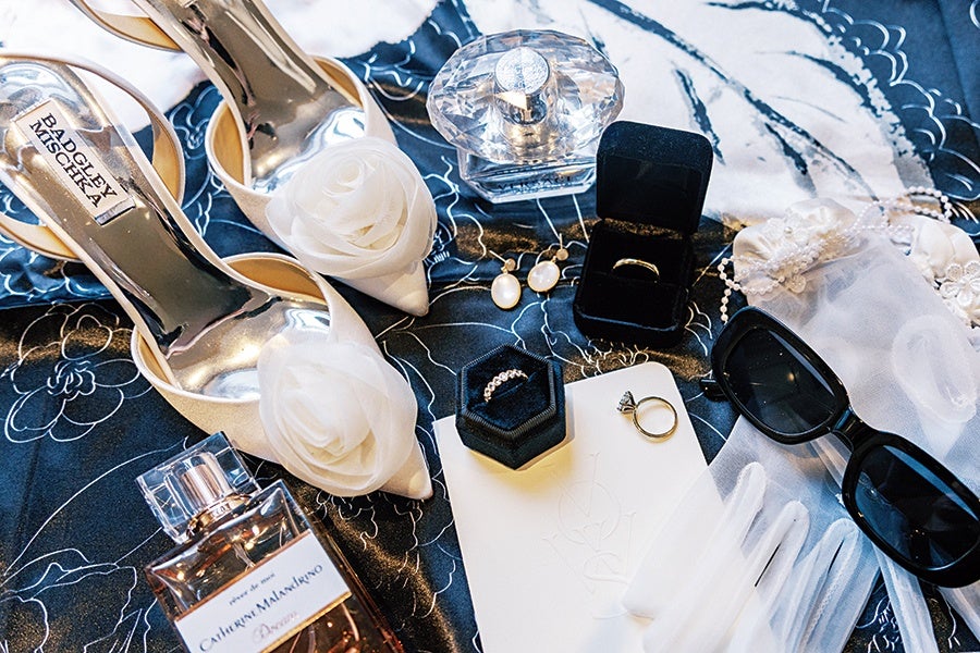 White bridal shoes with floral decorations, pearl earrings, two rings in black ring boxes, a diamond engagement ring, a pair of black sunglasses, white gloves, a pearl bracelet, and two perfume bottles are arranged on a dark floral fabric.