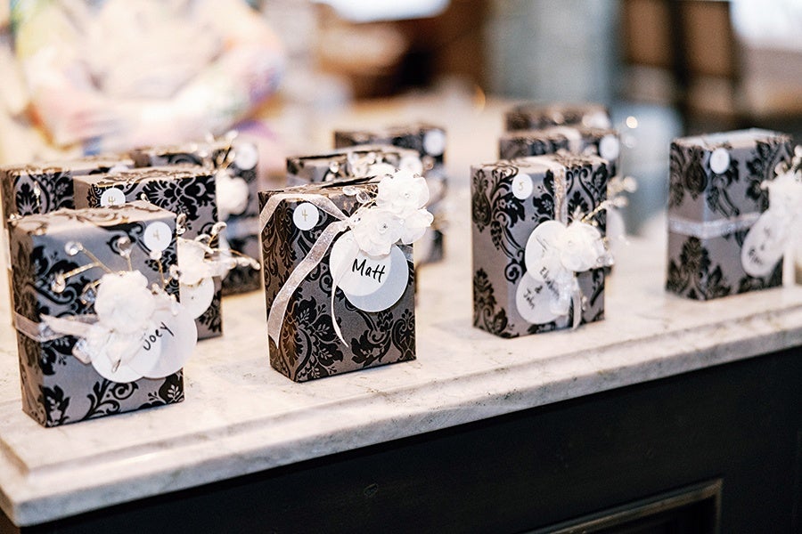Small rectangular gift boxes wrapped in black and silver patterned paper, each adorned with a white ribbon, a white flower decoration, and a round white name tag with handwritten names like "Matt" and "Joey," arranged on a marble surface.