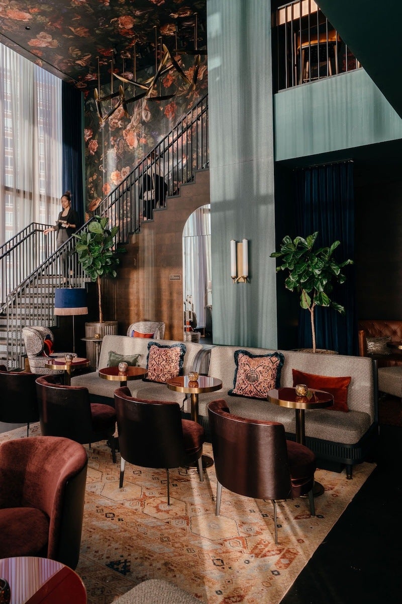 A stylish lounge area with a mix of seating including beige sofas adorned with patterned and solid-colored cushions, dark brown leather chairs, and a plush maroon armchair. Small round tables with gold bases are placed in front of the seating. The space features a patterned rug, large potted plants, and a staircase with a black railing leading to an upper level. The walls and ceiling near the staircase are decorated with a dark floral pattern, and tall windows with sheer curtains allow natural light to fill the room.