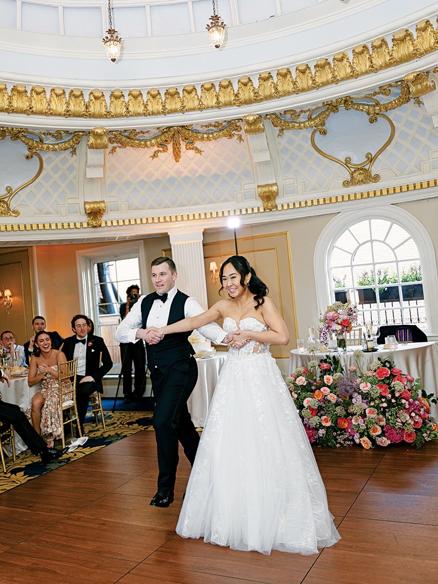 A bride and groom are dancing together on a wooden dance floor in an elegant room with ornate gold and white decor. The bride is wearing a strapless white wedding gown with floral details, and the groom is dressed in a black tuxedo with a white shirt and black bow tie. Behind them, guests seated at tables are watching and smiling. A large floral arrangement with pink, red, and purple flowers is placed near a round table covered with a white tablecloth. The room features a large arched window and decorative molding with gold accents.