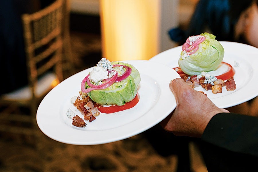 Two plates each with a wedge salad consisting of a wedge of iceberg lettuce topped with blue cheese, pickled red onions, and bacon bits, served on a bed of red dressing. A person is holding the plates.
