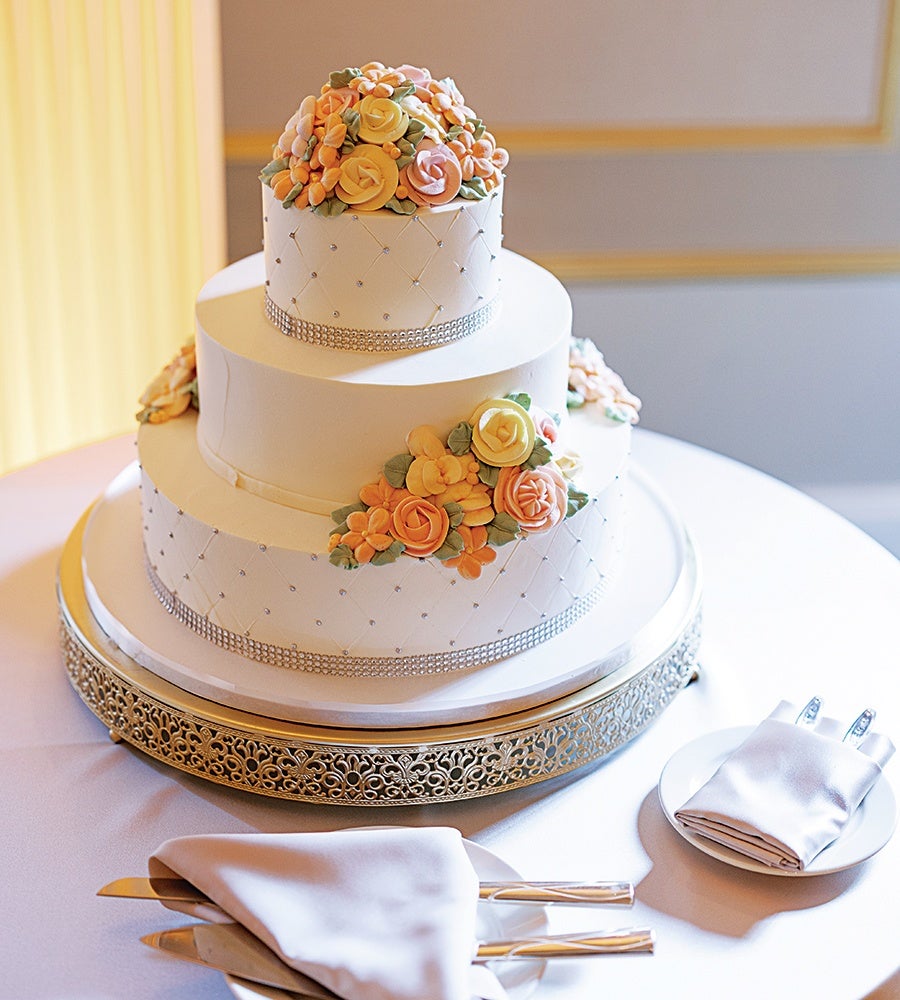 Three-tier white cake decorated with pastel yellow, orange, and peach buttercream flowers and green leaves. The cake features a quilted pattern with small silver beads and is accented with a silver rhinestone band around the base of each tier. It is displayed on an ornate silver cake stand on a white tablecloth, with folded white napkins and silver cake-cutting utensils nearby.