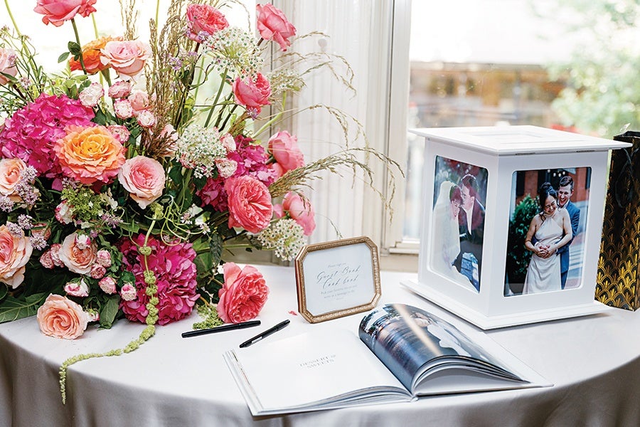 A wedding display on a round table with a white tablecloth, featuring a large floral arrangement of pink, peach, and orange roses and other flowers, a small framed sign, an open photo album, and a white photo lantern with pictures of a couple on its sides.