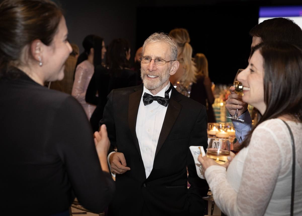 A man wearing a black tuxedo with a white shirt and black bow tie is smiling and engaging in conversation with two women. One woman on the left is wearing a black outfit, and the other woman on the right is wearing a white lace top and holding a glass of white wine. The background shows other people and a softly lit event setting.