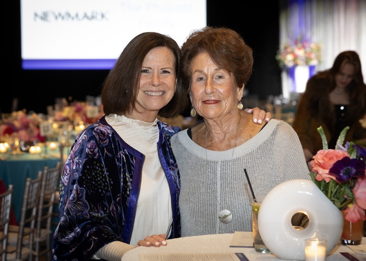 Two women are sitting closely together at a table, smiling at the camera. The woman on the left has shoulder-length brown hair and is wearing a white blouse with a dark blue floral-patterned jacket. The woman on the right has short curly brown hair and is wearing a light gray knit sweater with dangling earrings. On the table in front of them are a white decorative object, a glass with a straw, a lit candle, and a bouquet of pink and purple flowers. The background shows a softly lit event space with tables, chairs, and floral arrangements.