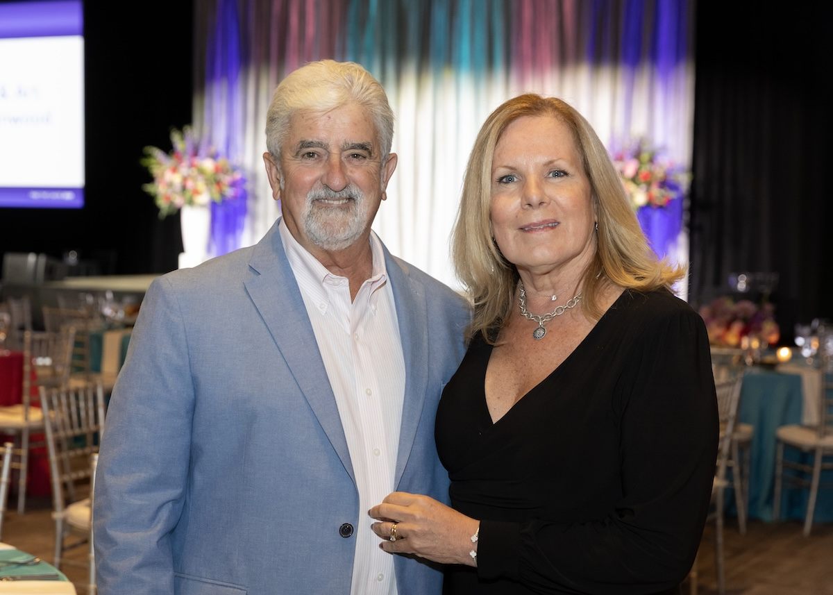 A man and a woman standing close together indoors at an event. The man has white hair and a beard, wearing a light blue blazer over a white shirt. The woman has blonde hair, wearing a black dress and a silver necklace. The background shows tables with chairs and floral arrangements, with colorful draped curtains.