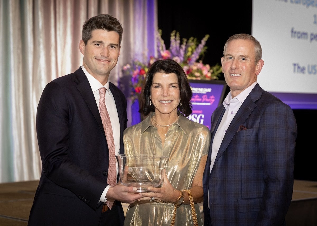 Three people dressed in formal attire stand together, smiling. The man on the left wears a dark suit with a white shirt and a pink tie. The woman in the center wears a shiny gold dress and holds a clear glass award. The man on the right wears a dark blue plaid suit with a light-colored shirt. Behind them is a blurred background with flowers and a screen displaying text.
