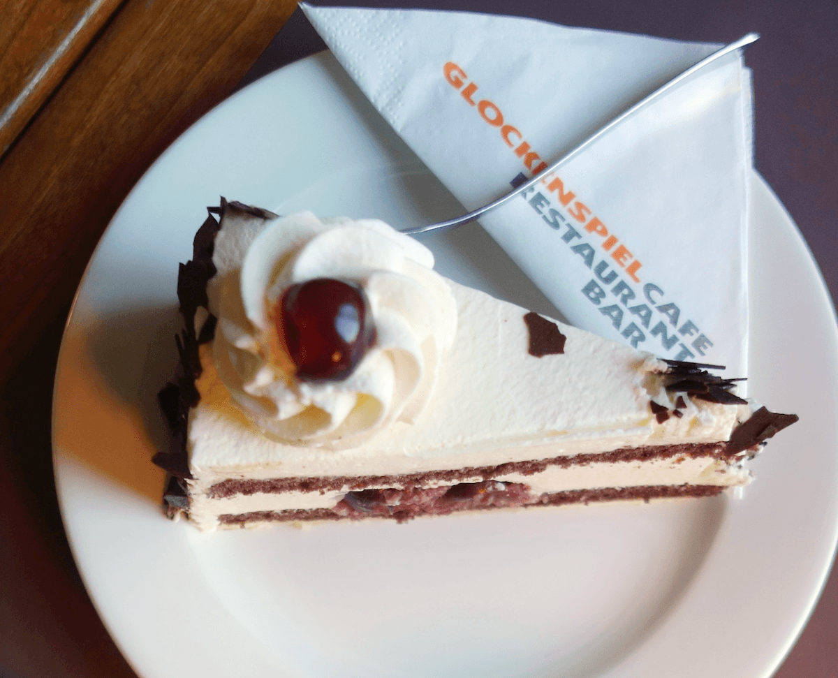 A slice of Black Forest cake with layers of chocolate sponge, cream, and cherry filling, topped with whipped cream and a single cherry. The cake is served on a white plate with a folded napkin and a fork. The napkin has the text "GLOCKENSPIEL CAFE RESTAURANT BAR" printed on it.