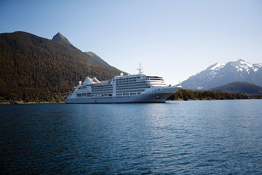 A large white cruise ship sailing on calm blue water with forested mountains on the left and snow-capped mountains on the right under a clear blue sky.