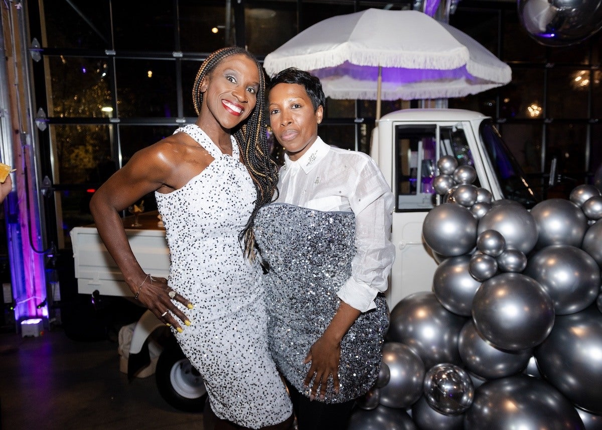 Two women posing together indoors at an event. The woman on the left is wearing a white sleeveless dress with black speckles and has long braided hair. She is smiling broadly with her hand on her hip. The woman on the right is wearing a white shirt with a silver sequined overlay and has short hair. Behind them is a white vehicle with a white umbrella and a large cluster of metallic silver balloons. The background features large windows and purple lighting.