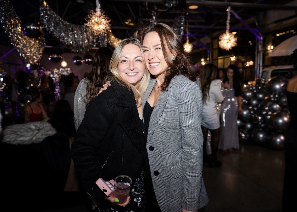 Two women smiling and posing closely together at a festive indoor event. One woman has blonde hair, wears a black jacket, and holds a drink and a pink phone. The other woman has brown hair, wears a gray plaid blazer over a black top, and is smiling broadly. The background features decorative silver balloons, hanging lights, and other people dressed in party attire.