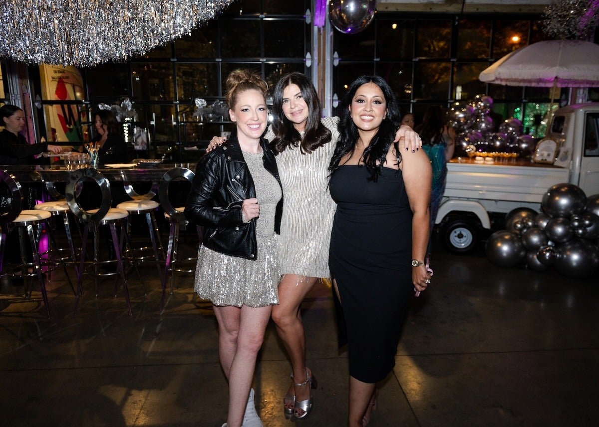 Three women posing together indoors at a party or event. The woman on the left is wearing a sparkly silver dress with a black leather jacket and white shoes, with her hair tied up. The woman in the middle is wearing a short, silver, sequined dress and silver high heels, with her dark hair down. The woman on the right is wearing a fitted black dress with a slit and a watch on her left wrist, with long dark hair. The background features a bar area with stools, silver balloons, and a white truck decorated with more silver balloons.