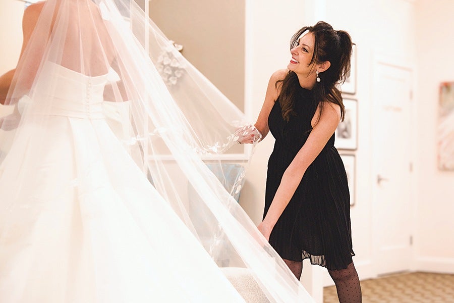 A woman in a sleeveless black dress with sheer polka-dot tights is smiling and holding the veil of a bride wearing a white wedding gown with a fitted bodice and button details on the back. The scene appears to be indoors with soft lighting and framed pictures on the wall.