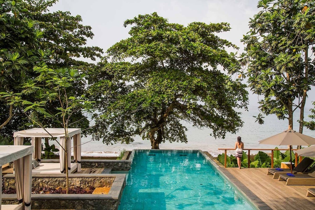A rectangular outdoor swimming pool with clear blue water is surrounded by a wooden deck and stone borders. On the left side, there are two white canopy beds with curtains, and on the right side, there are lounge chairs with beige umbrellas. A large leafy tree stands at the far end of the pool, overlooking a body of water in the background. A person wrapped in a towel is standing near the wooden railing, facing the water. The scene is lush with greenery and has a serene, tropical atmosphere.