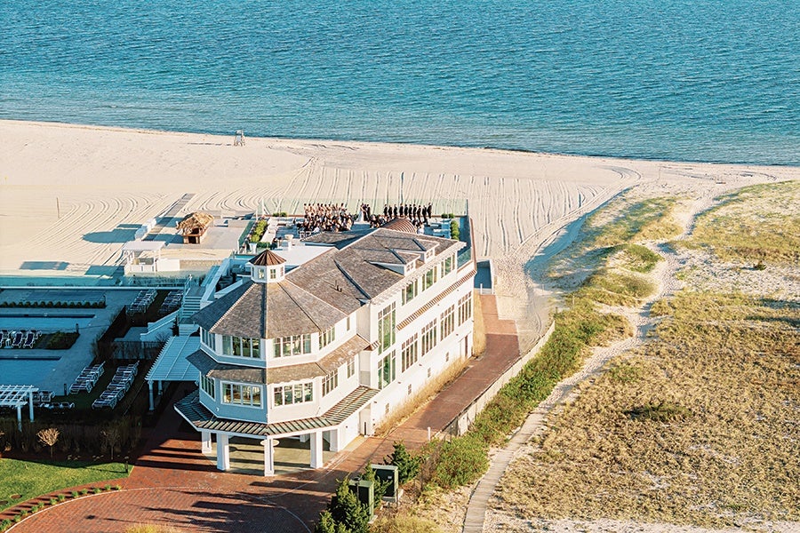 A large, multi-story white building with a gray roof is situated near a sandy beach and ocean. The building has many windows and a covered entrance supported by columns. There is a paved walkway and some greenery around the building, with sand dunes and a wooden path leading to the beach nearby. The ocean is calm and blue in the background.