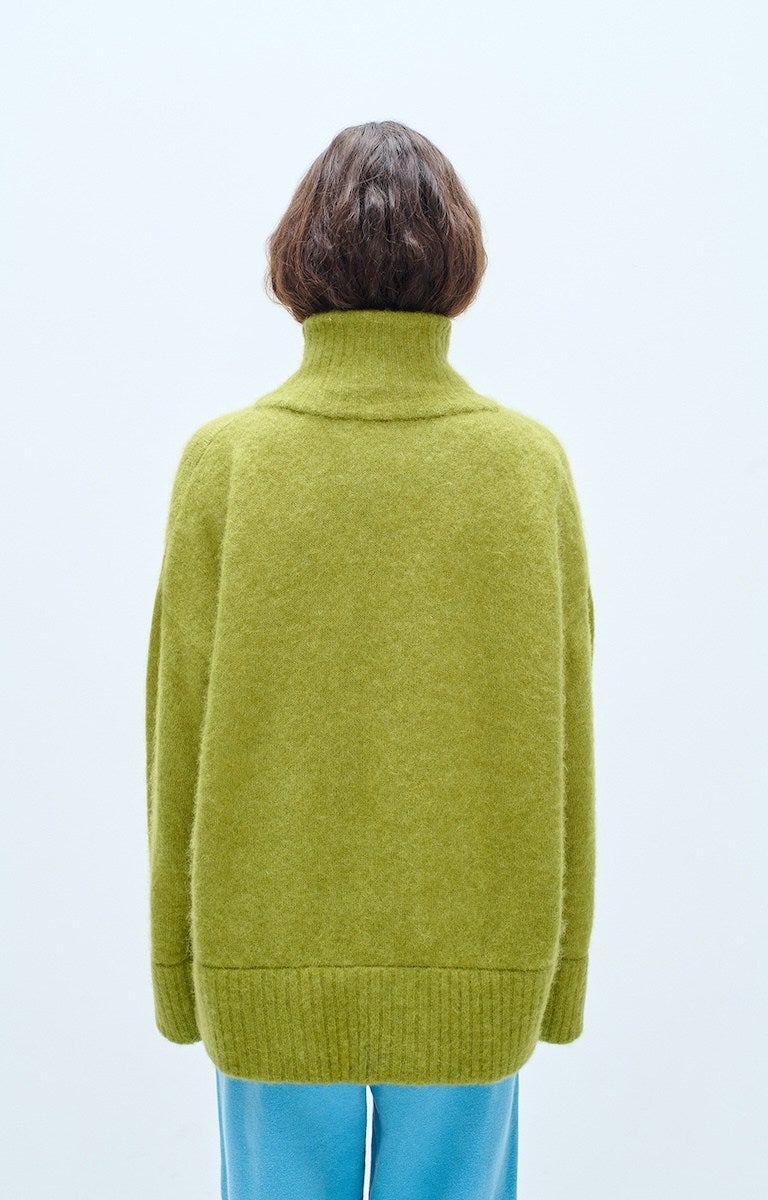 A person with short brown hair wearing a loose-fitting, olive green turtleneck sweater and light blue pants, shown from the back against a plain light background.