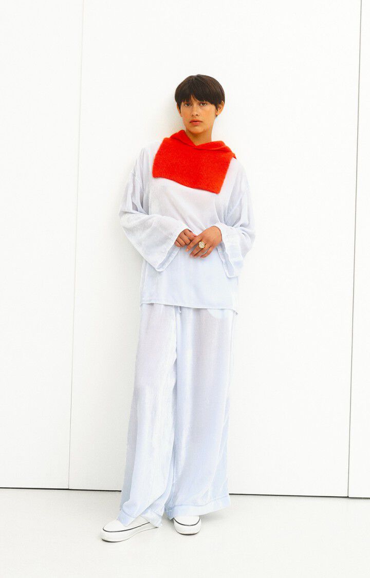 A person wearing a loose-fitting, light blue outfit consisting of a long-sleeve top with wide sleeves and wide-leg pants. They have a bright red knitted scarf draped around their neck and are wearing white slip-on shoes. The background is plain white.