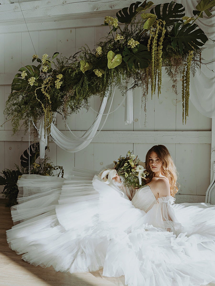 A woman in a voluminous white wedding dress with off-the-shoulder sleeves is sitting on the floor, holding a bouquet of flowers. Behind her is a large floral arrangement with green leaves and hanging yellow-green flowers, draped with white fabric and pearl-like strands, set against a white wooden wall.
