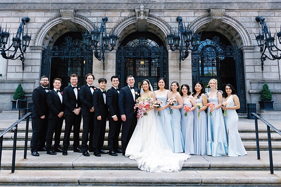 A wedding party posed on stone steps in front of an ornate building with arched doorways and black wrought iron gates. The group includes six groomsmen in black tuxedos and bow ties, the groom in a black tuxedo, the bride in a white lace wedding gown holding a large bouquet of pink and white flowers, and six bridesmaids wearing light blue dresses, each holding smaller bouquets of pink and white flowers. The setting features large black lantern-style lamps on either side of the steps.