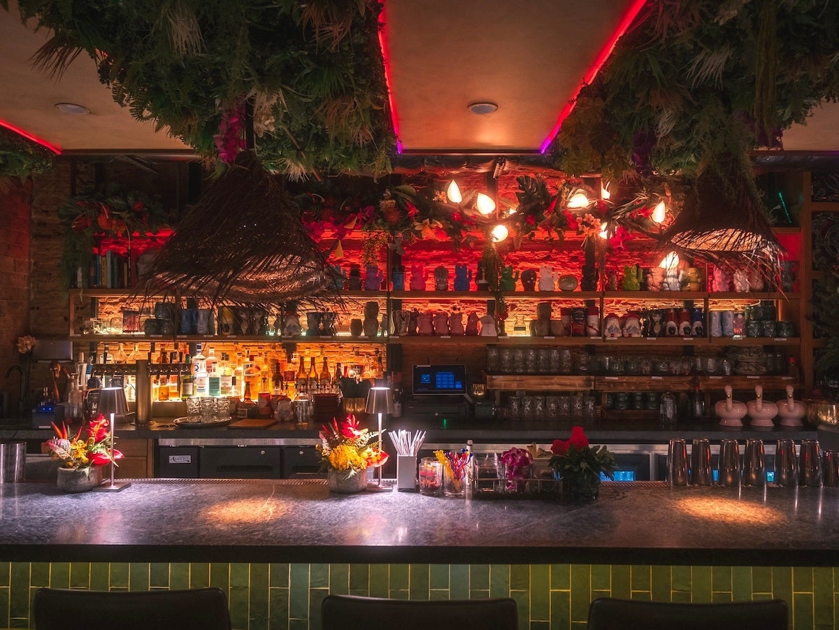 A dimly lit bar with a stone countertop and green tiled front. The back wall features shelves filled with various bottles, glassware, and tiki-style mugs, illuminated by warm orange and red lighting. Two large woven pendant lamps hang from the ceiling, surrounded by hanging greenery and floral decorations. Small floral arrangements and bar tools are placed on the counter.