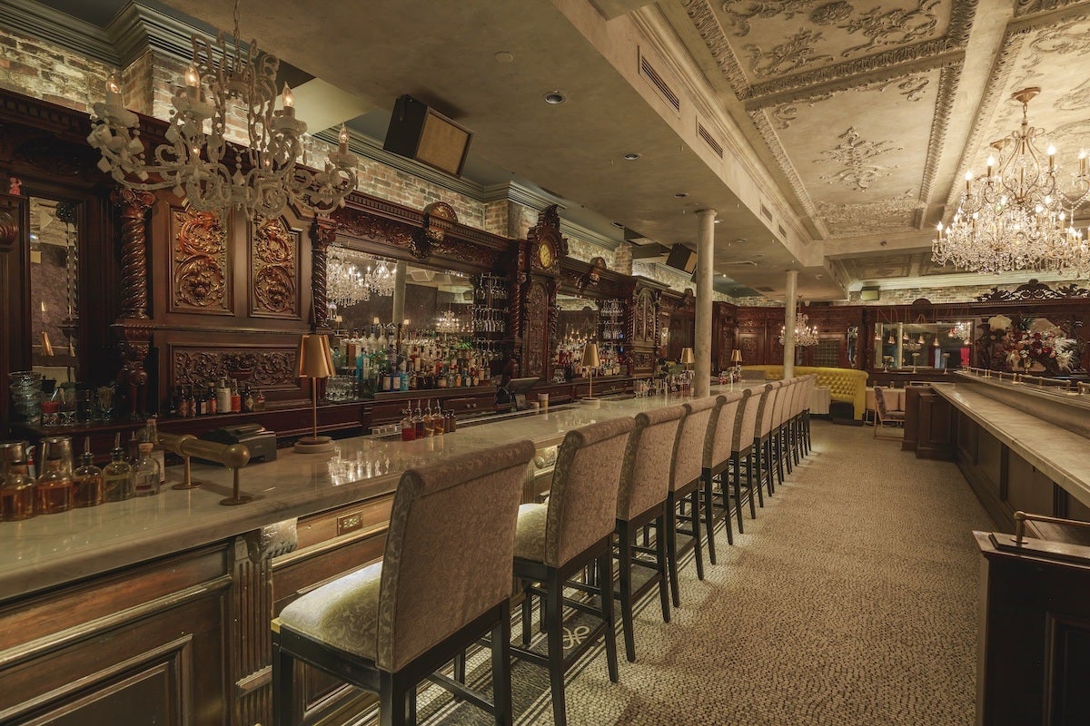 A long, elegant bar with a marble countertop and a row of upholstered bar stools featuring patterned fabric. The back wall is adorned with intricately carved dark wood panels and large mirrors, reflecting crystal chandeliers hanging from a decorative, ornate ceiling. The bar is stocked with various bottles and glassware, and the floor is covered with a textured carpet. The overall ambiance is luxurious and vintage.
