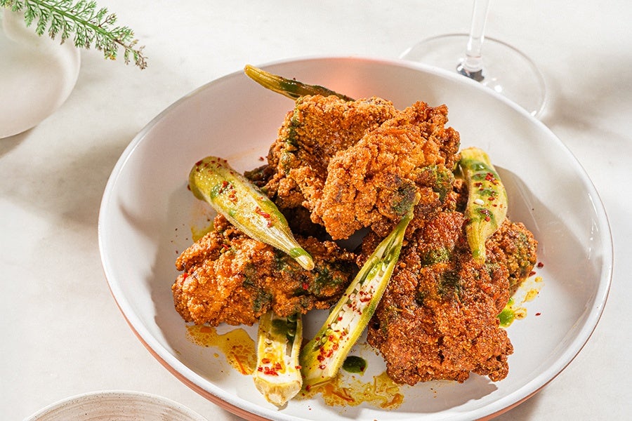 A white bowl contains several pieces of crispy fried chicken garnished with green herbs and spices. Alongside the chicken are whole fried okra pods sprinkled with red chili flakes. The dish is presented on a light-colored surface with a glass stemware partially visible in the background.