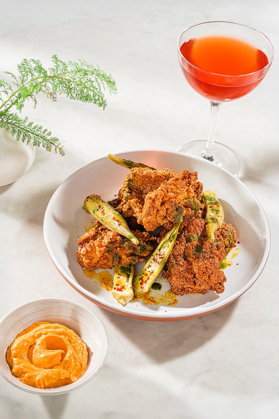 A white bowl contains crispy fried chicken pieces garnished with sliced okra and sprinkled with red chili flakes and green herbs. Next to the bowl is a small dish of creamy orange dipping sauce. A glass of bright red-orange cocktail is placed behind the bowl. A small green plant in a white vase is visible in the background on a light-colored surface.