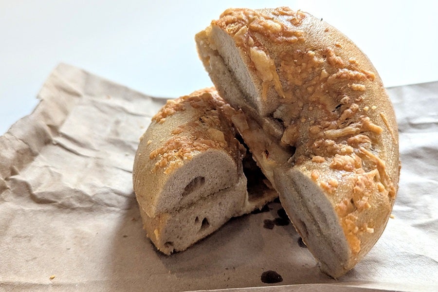 A sliced bagel with a golden-brown crust topped with melted cheese, resting on a crumpled piece of brown paper. The interior of the bagel appears soft and slightly dense.