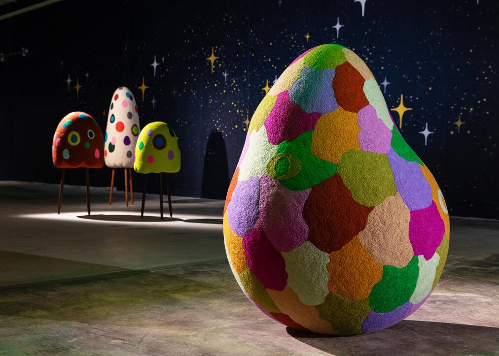 A large, egg-shaped sculpture covered in a patchwork of colorful, textured fabric pieces in shades of green, purple, orange, pink, and beige stands prominently in the foreground. In the background, three smaller, similarly shaped sculptures with bright polka dots on red, white, and yellow bases are elevated on thin legs. The setting appears to be an indoor space with a dark wall decorated with star-like patterns.