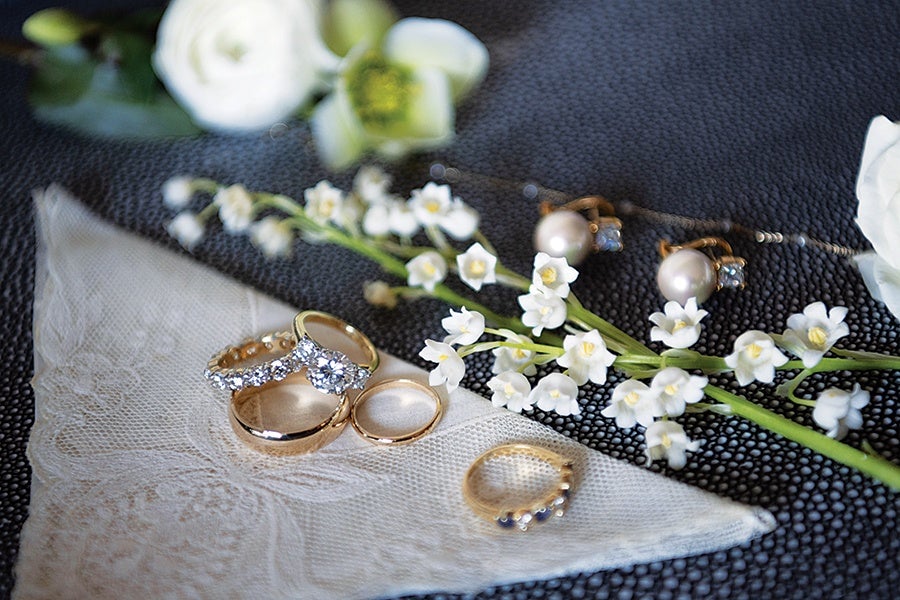 A collection of gold rings, including one with a large central diamond and another with smaller diamonds, is displayed on a white lace handkerchief. Nearby, a pair of pearl earrings with small blue stones and a delicate chain are placed next to a sprig of small white flowers on a textured dark surface. White and green flowers are blurred in the background.