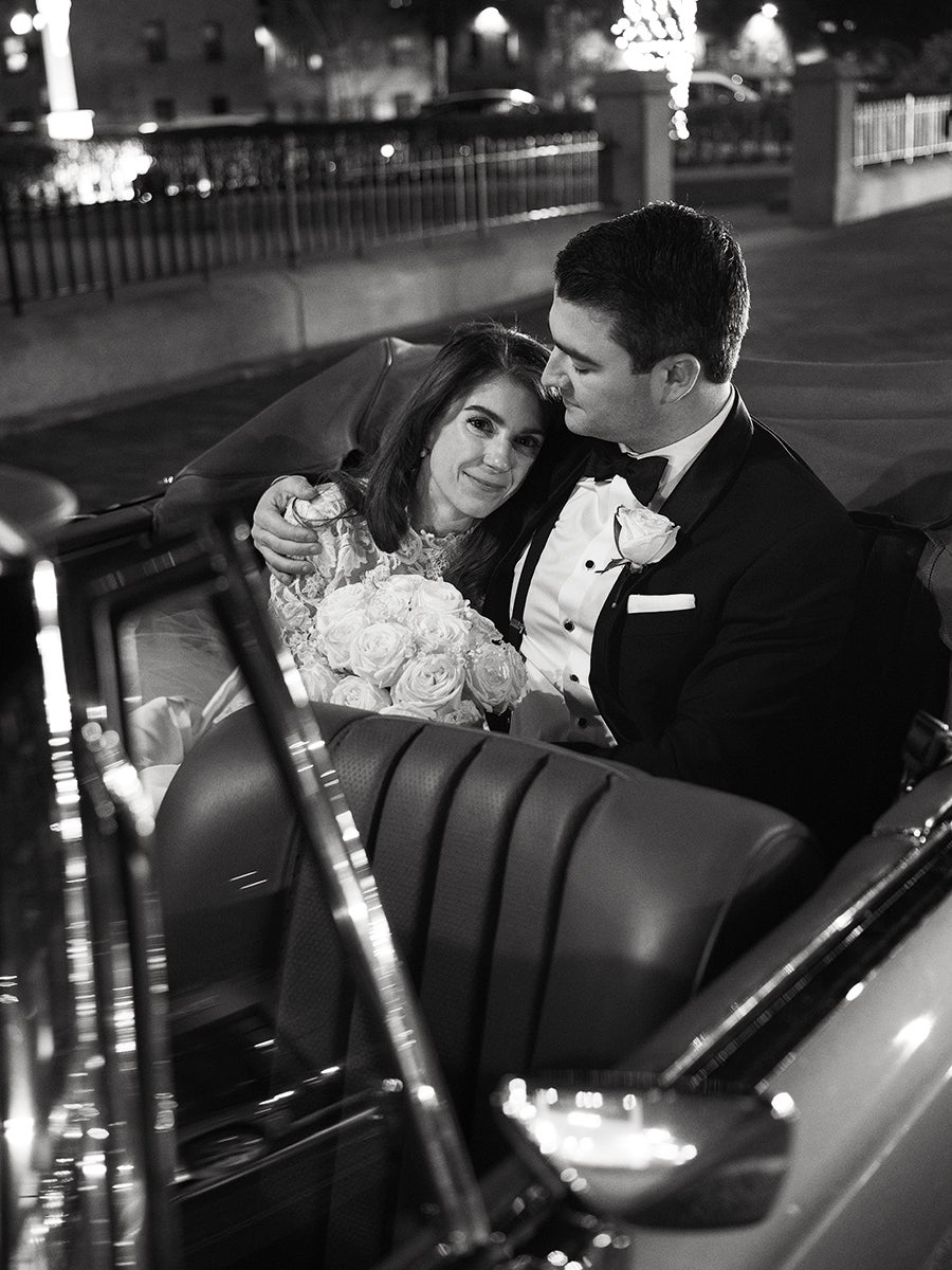 A black and white photo of a bride and groom sitting closely together in the backseat of a vintage car at night. The bride is holding a bouquet of roses and leaning into the groom, who is wearing a tuxedo with a bow tie and a rose boutonniere. The groom has his arm around the bride, and they both appear content and affectionate. The background shows a dimly lit street with some lights and a fence.