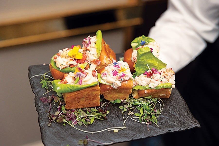 Five pieces of gourmet finger food served on a black slate platter, each consisting of a square of bread topped with lettuce, a creamy seafood salad, and garnished with colorful edible flowers and microgreens.