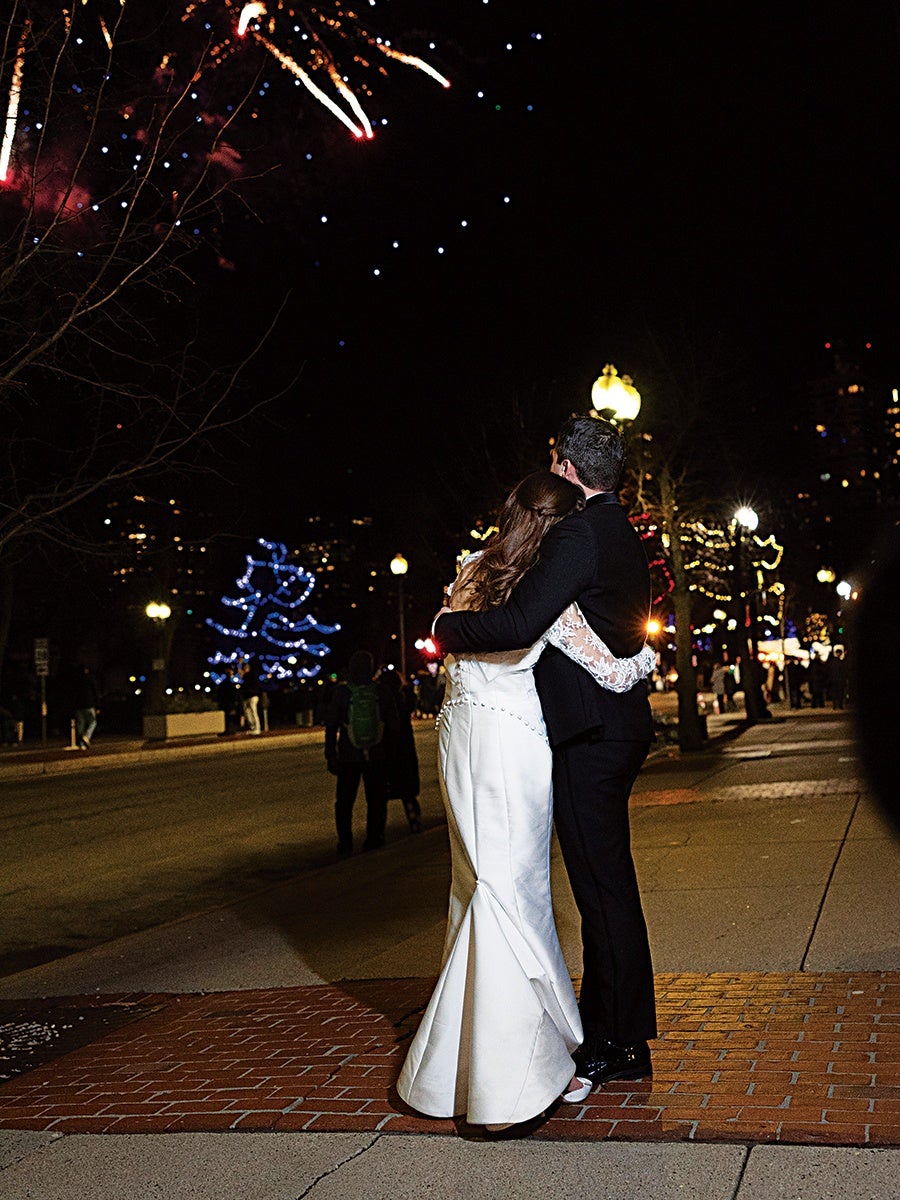 A couple dressed in formal wedding attire embraces on a city sidewalk at night. The bride wears a white gown with lace sleeves, and the groom is in a black suit. Fireworks light up the dark sky above, and festive lights decorate the trees and street lamps in the background.