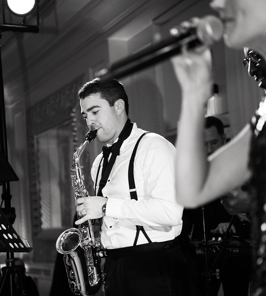 A man dressed in a white shirt with black suspenders and a bow tie is playing a saxophone. In the foreground, a person is holding a microphone close to their mouth, partially out of focus. The scene appears to be indoors with a decorative wall and lighting equipment visible in the background. The image is in black and white.