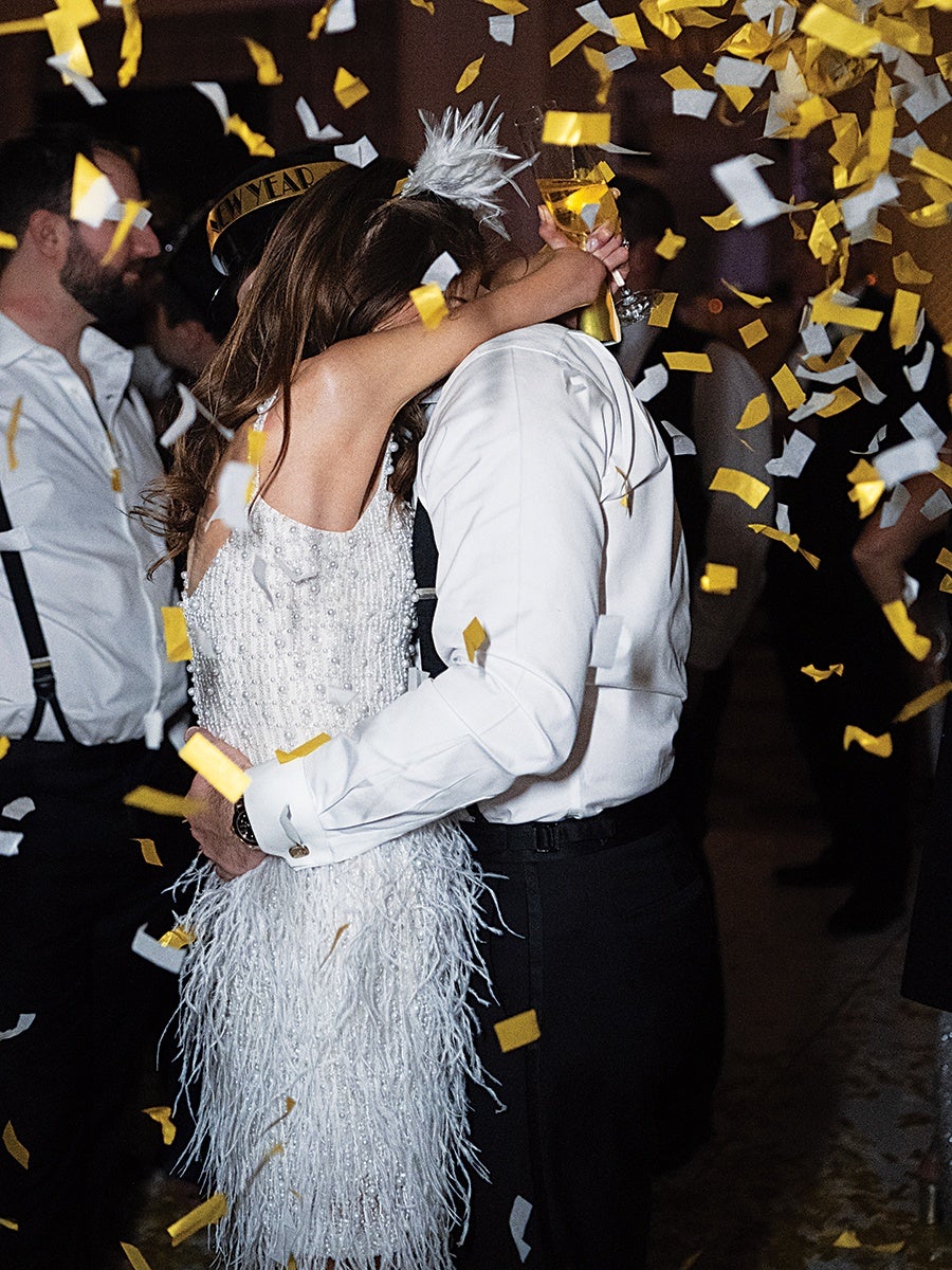 A couple embracing and kissing at a celebration with gold and white confetti falling around them. The woman is wearing a white dress adorned with beads and feathers, and a white feathered headpiece. The man is dressed in a white shirt and black pants, holding a glass of champagne. Other people in festive attire are visible in the background.