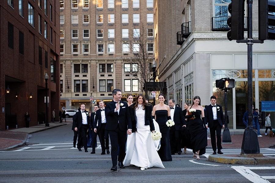 A wedding party crossing a city street, with the bride in a white gown holding a bouquet, the groom in a black tuxedo, and bridesmaids and groomsmen dressed in formal black attire. The background features tall buildings and a traffic light showing a red hand signal.