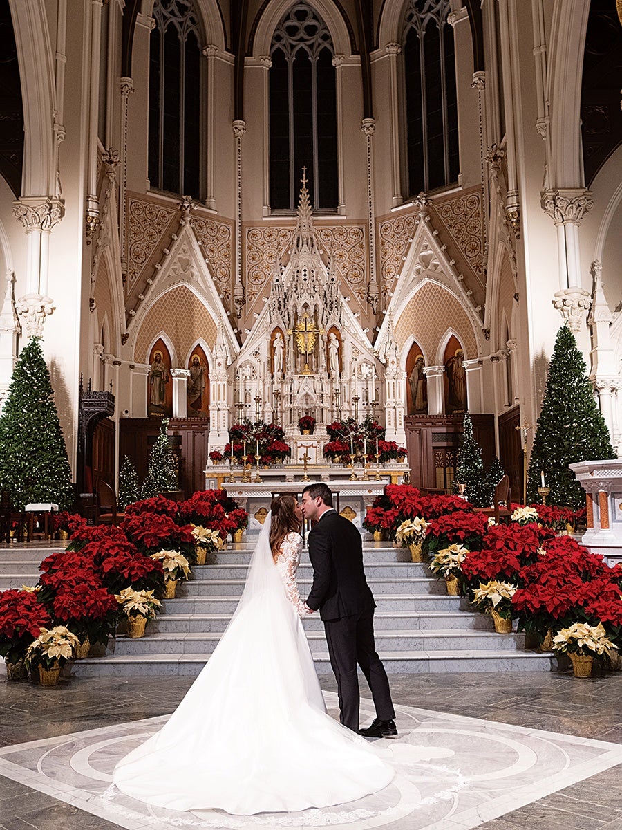 A bride and groom stand at the altar of a grand church, sharing a kiss. The bride wears a long white gown with a lace bodice and a flowing train, while the groom is dressed in a black suit. The altar is ornately decorated with red and white poinsettias, tall Christmas trees, and intricate architectural details, including a large, ornate white structure with religious statues and a gold cross. The setting is elegant and festive, with high arched windows and detailed wall patterns.