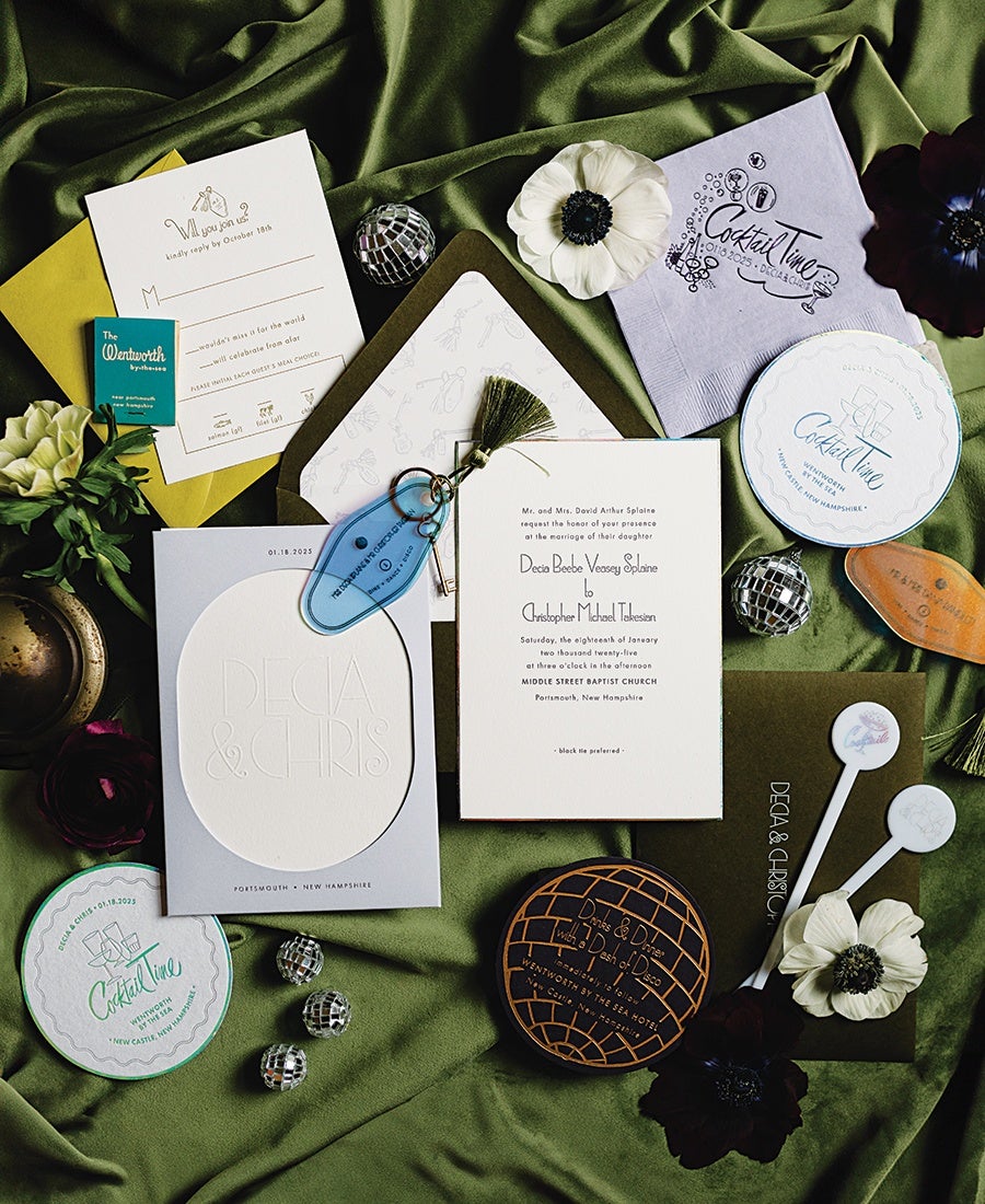 A wedding invitation suite displayed on green fabric, featuring white and olive green cards with elegant typography. The main invitation card includes the names Decia Beebe Veasey Splaine and Christopher Michael Takesian, with details of the wedding date and location in Portsmouth, New Hampshire. Surrounding the invitations are cocktail napkins and coasters labeled "Cocktail Time," small disco ball decorations, white and dark purple flowers, a blue key tag, and two white drink stirrers with round tops. The overall arrangement is stylish and festive.