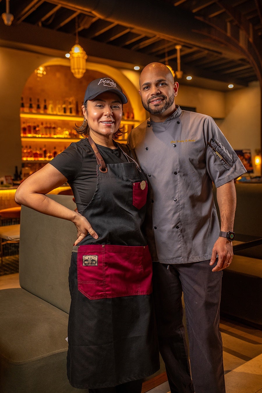 A man and a woman standing together in a warmly lit restaurant setting. The woman is wearing a black and red apron over a black shirt and a black cap, while the man is dressed in a gray chef's coat and dark pants, with a smartwatch on his left wrist. Both are smiling and appear to be staff or chefs at the restaurant. The background features shelves with bottles and warm ambient lighting.