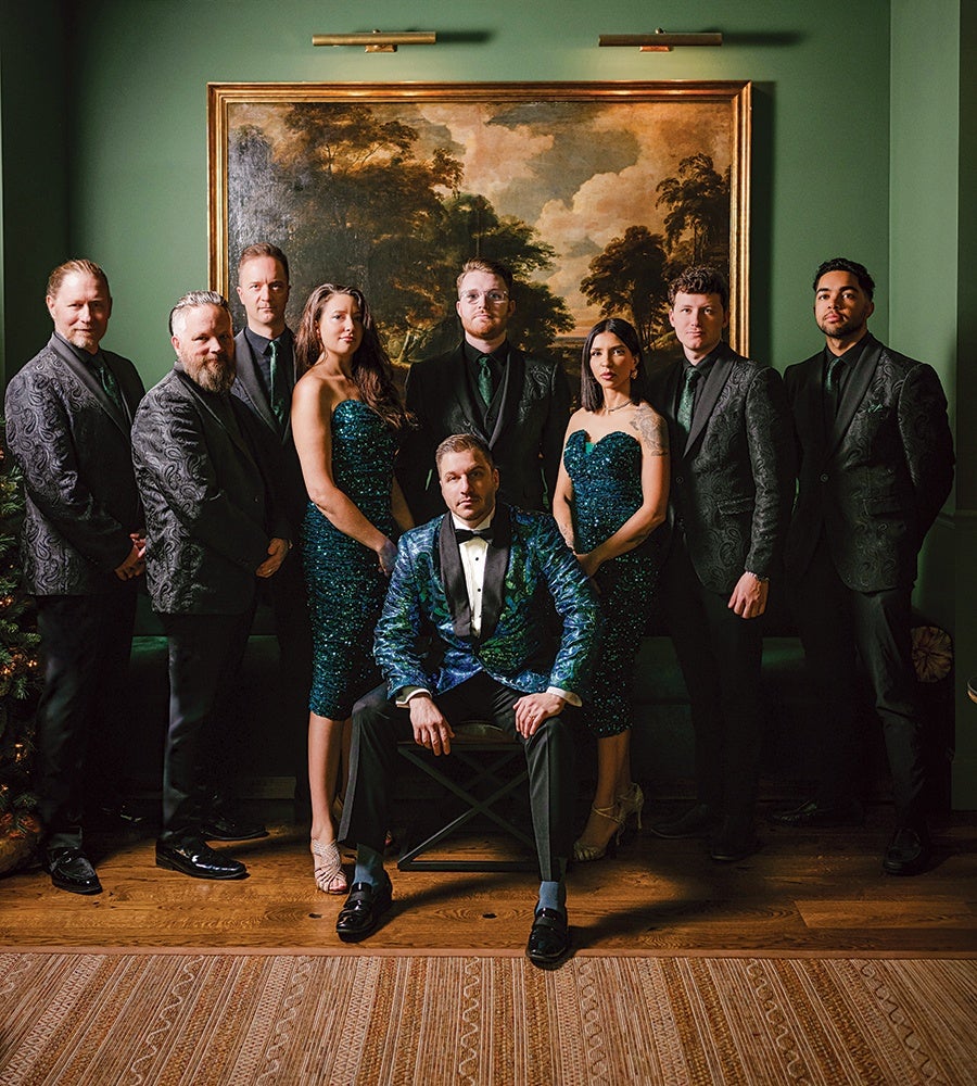 A formal group portrait of nine people dressed in elegant evening attire. Eight individuals stand in a row, with two women wearing sparkly teal dresses and six men in black suits with patterned jackets and dark green ties. One man sits in front on a chair, wearing a shiny blue and green patterned tuxedo jacket with black pants, a white shirt, and a black bow tie. The background features a large framed landscape painting on a green wall, and the floor is wooden with a patterned rug in the foreground.