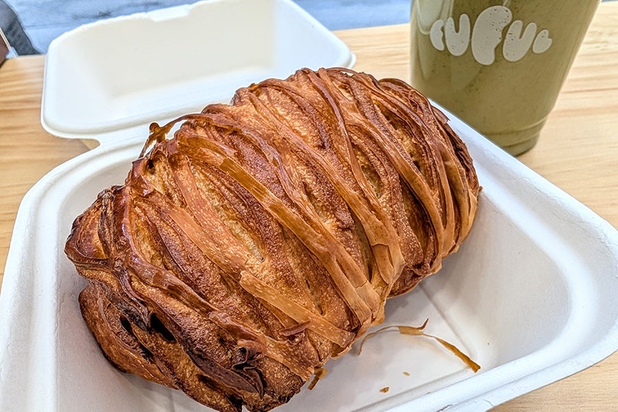 A golden-brown braided pastry with a flaky texture sits inside a white takeout container on a wooden table. In the background, there is a green cup with white text on it.