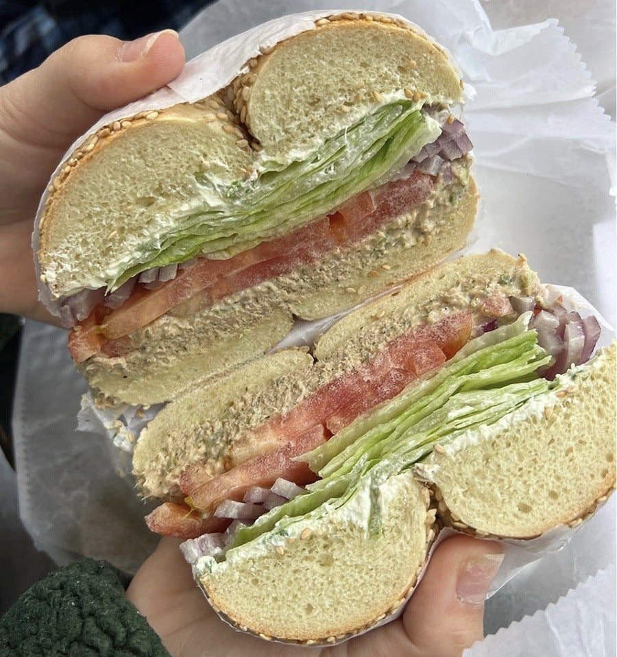 Sesame seed bagel sandwich cut in half, filled with layers of lettuce, tomato slices, chopped red onions, a creamy spread, and tuna. The sandwich is held with both hands and wrapped partially in white paper.
