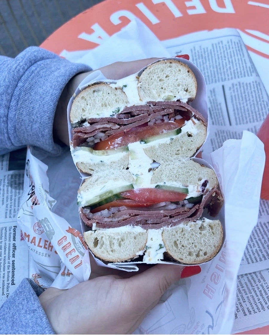 A sliced bagel sandwich held in two hands with red-painted nails, filled with layers of thinly sliced meat, tomato, cucumber, onions, and a generous amount of cream cheese. The sandwich is wrapped in deli paper, and the background includes a newspaper and an orange table surface.