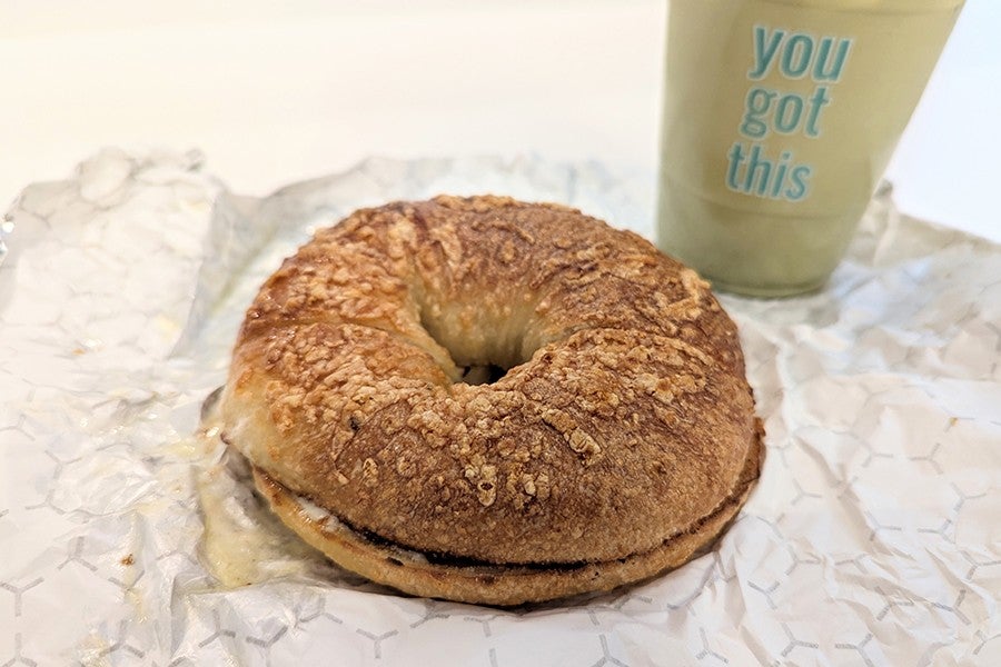 Toasted bagel sandwich with melted cheese on a patterned white wrapper, accompanied by a light green beverage in a cup that has the phrase "you got this" printed in blue.