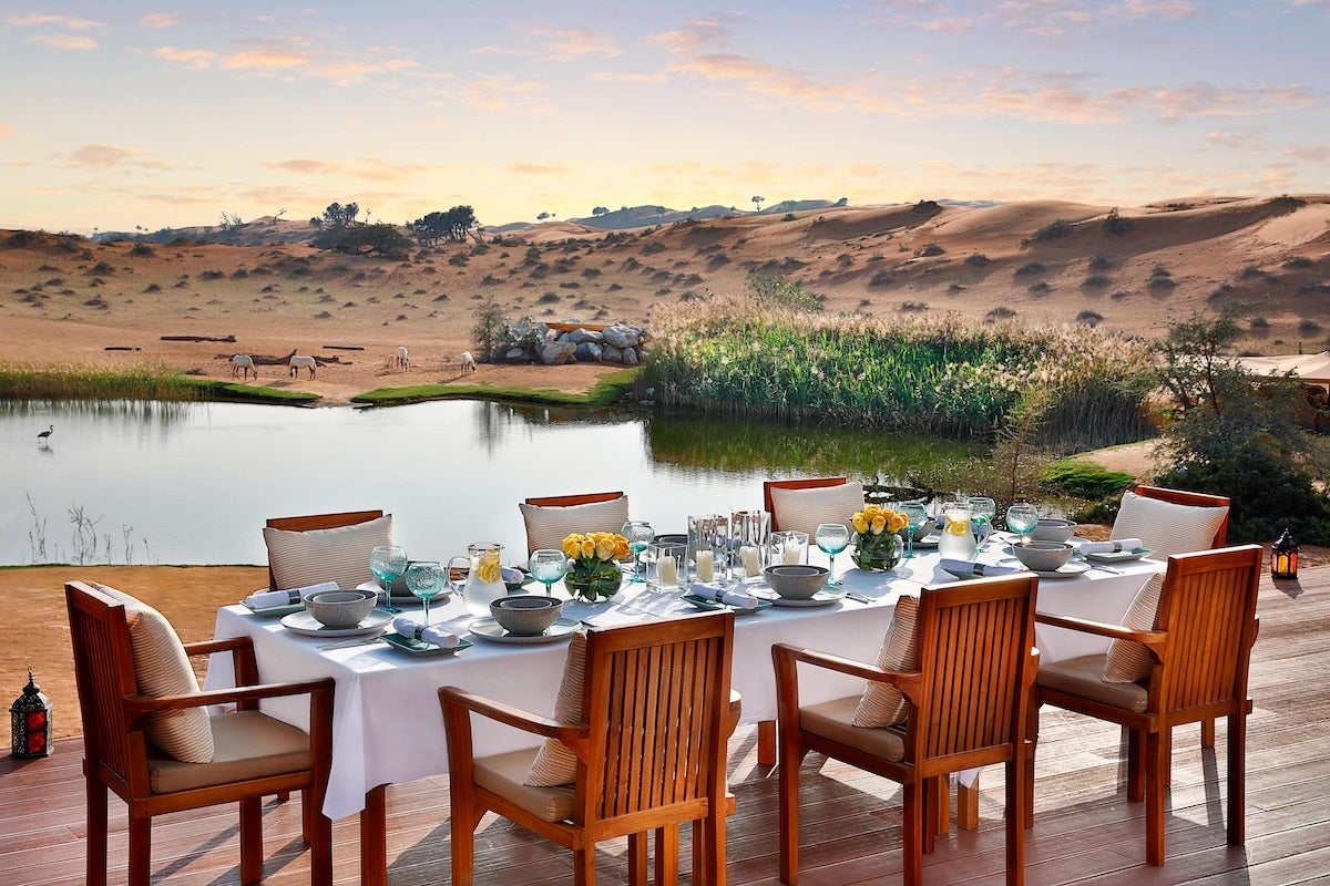 Outdoor dining table set with a white tablecloth, eight wooden chairs with cushions, glassware, bowls, pitchers of water with lemon, yellow flower centerpieces, and candles, overlooking a serene lake with reeds and a desert landscape with grazing animals in the background at sunset.