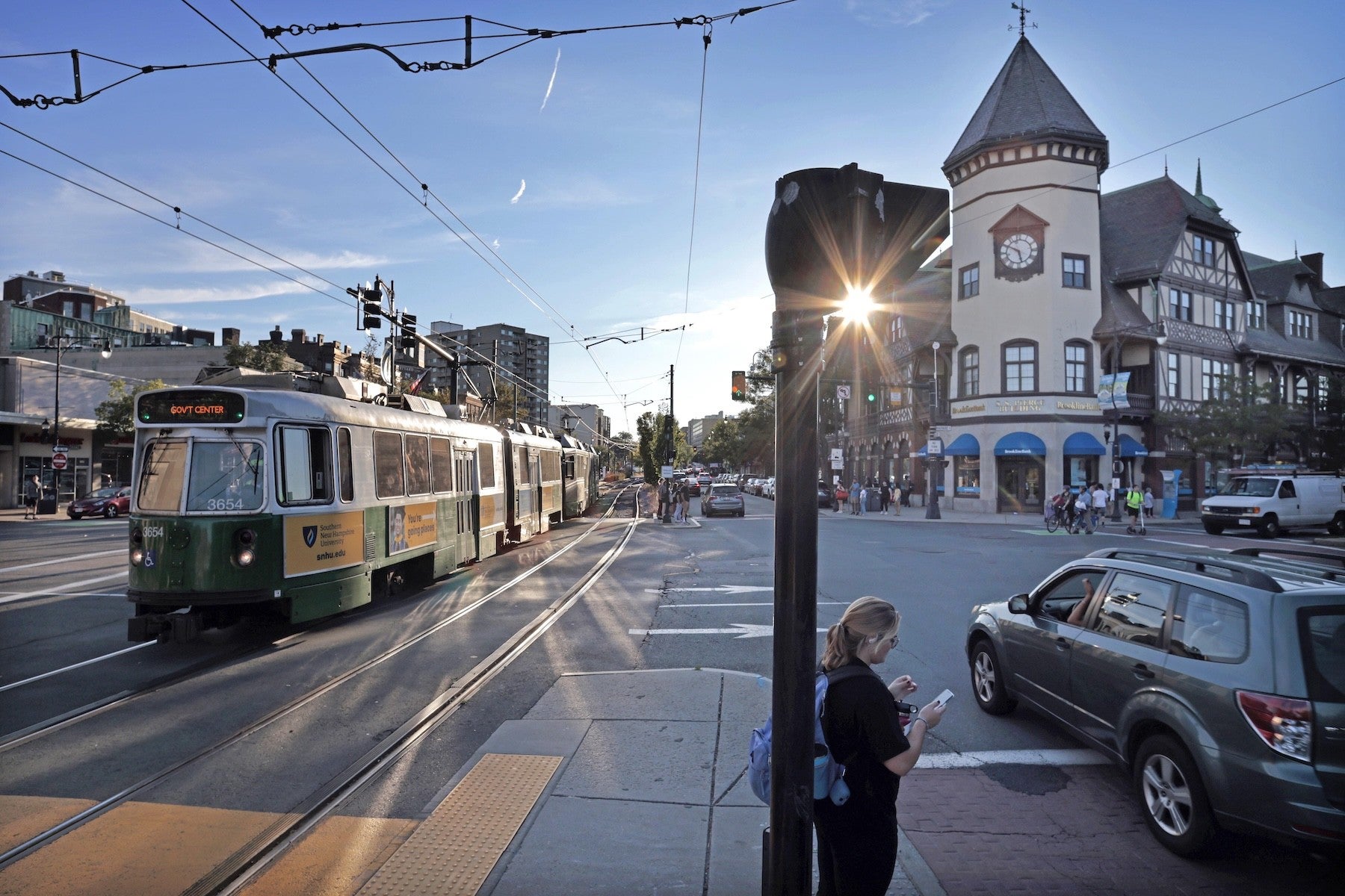 A green and white streetcar labeled "GOVT CENTER" travels on tracks along a city street. Overhead wires power the streetcar. A woman with a blue backpack stands on the sidewalk near a traffic light pole, looking at her phone. To the right, a dark green SUV waits at the intersection. In the background, a large building with a clock tower and Tudor-style architecture is visible, with people walking nearby. The sun is low, shining through the traffic light, casting long shadows.